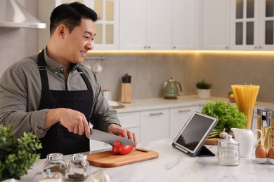 Photo of Cooking process. Man using tablet while cutting fresh bell pepper at countertop in kitchen
