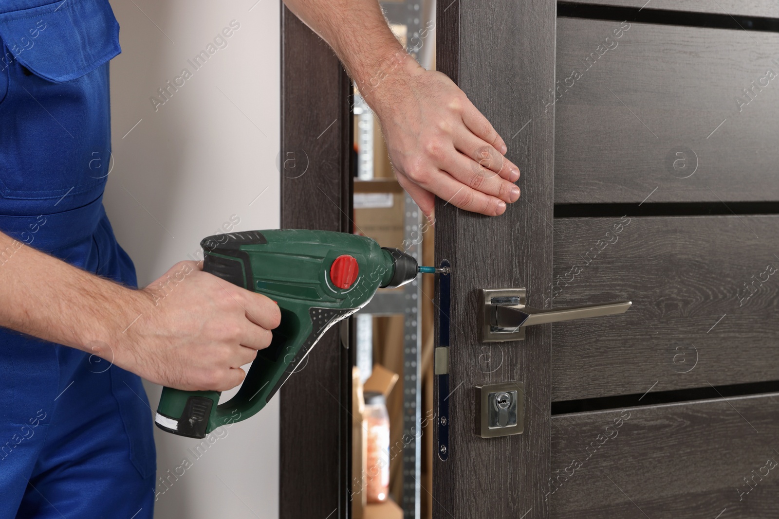 Photo of Worker in uniform with screw gun repairing door lock indoors, closeup