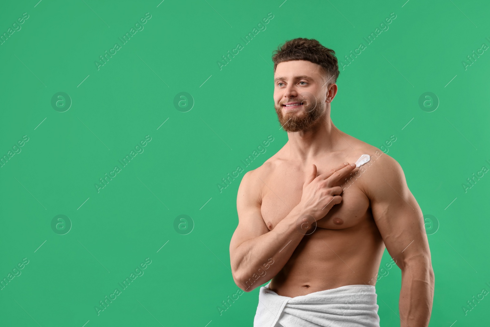 Photo of Handsome man applying moisturizing cream onto his shoulder on green background, space for text