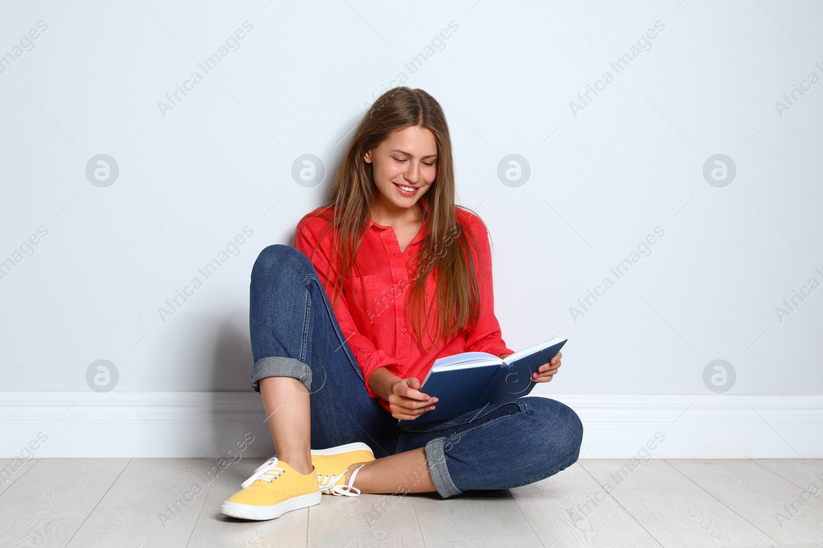 Photo of Beautiful young woman reading book on wooden floor near light wall