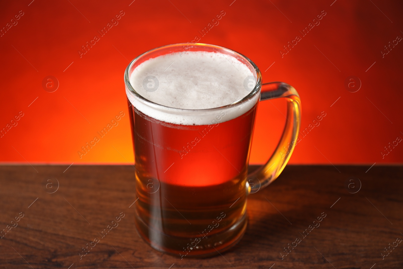 Photo of Mug with fresh beer on wooden table against red background, closeup