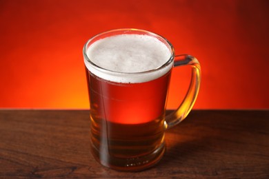 Photo of Mug with fresh beer on wooden table against red background, closeup