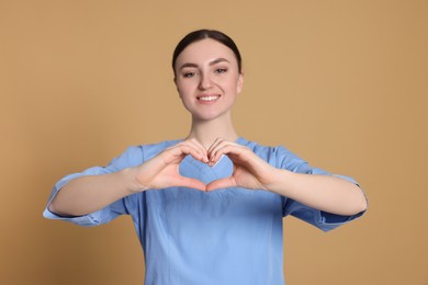 Photo of Nurse in medical uniform making heart with hands against light brown background, focus on palms