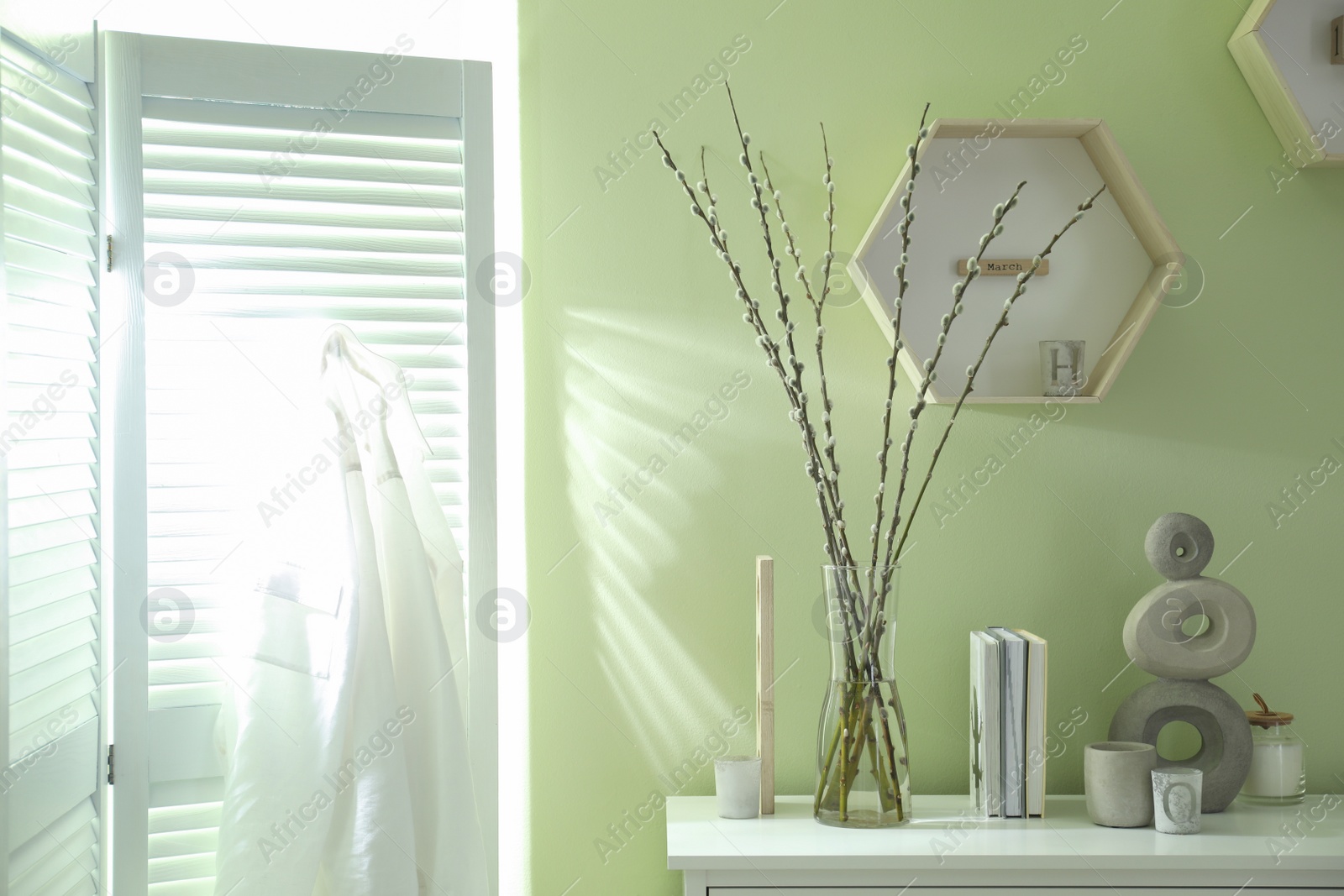 Photo of Glass vase with pussy willow tree branches and decor on white table near light green wall indoors