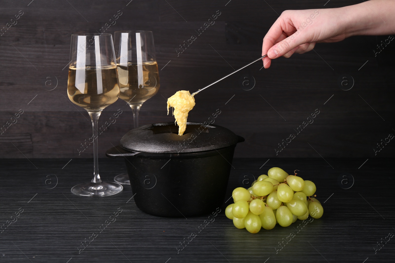 Photo of Woman dipping grape into fondue pot with melted cheese at black wooden table, closeup