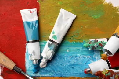 Photo of Tubes of colorful oil paints and spatula on canvas with abstract painting, flat lay
