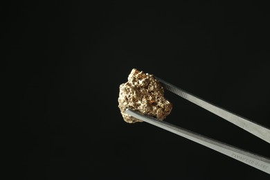 Tweezers with gold nugget against black background, closeup. Space for text