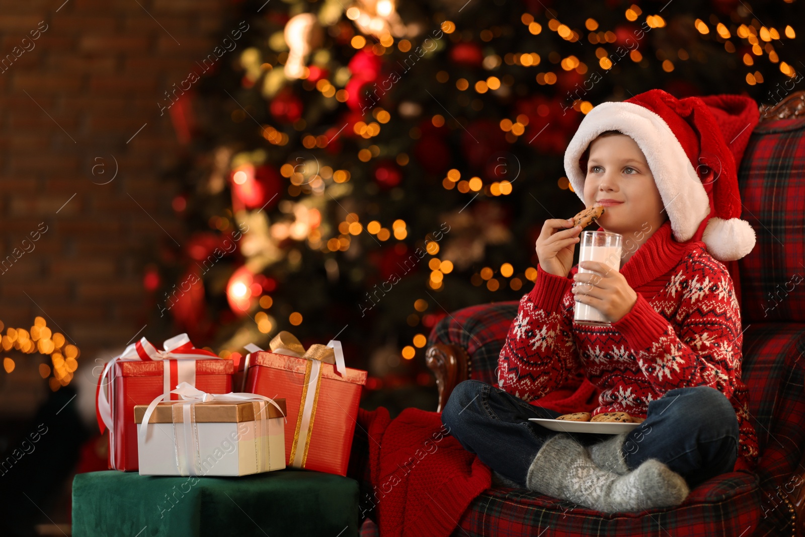 Photo of Little boy in Santa Claus cap with milk and cookies near Christmas gifts at home