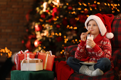 Photo of Little boy in Santa Claus cap with milk and cookies near Christmas gifts at home