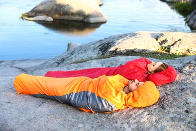 Young couple resting in sleeping bags on cliff near lake