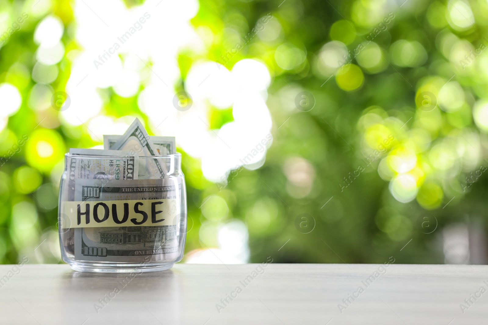 Photo of Glass jar with money and label HOUSE on table against blurred background. Space for text