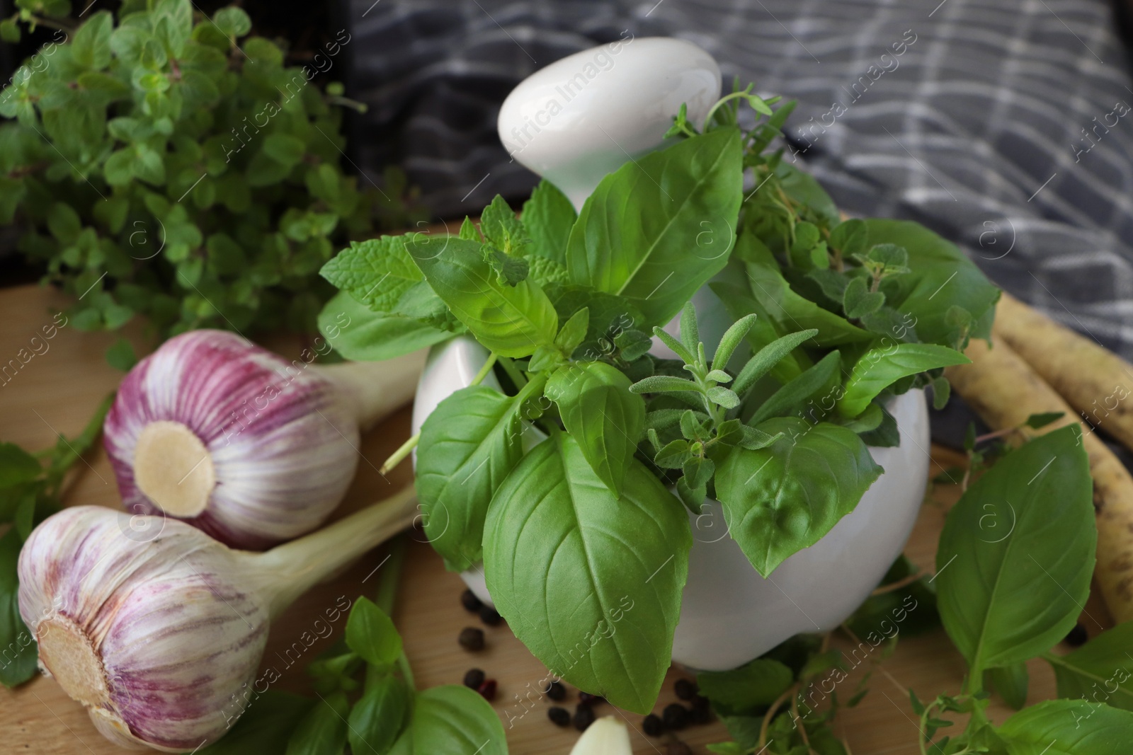 Photo of Mortar with different fresh herbs near garlic, horseradish roots and black peppercorns on wooden table, closeup