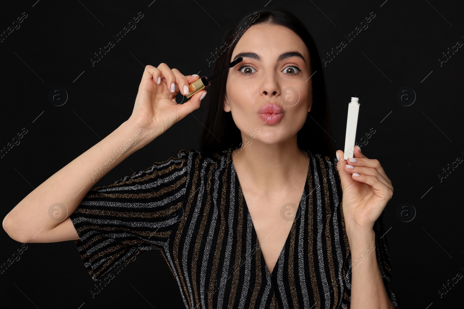 Photo of Beautiful young woman sending air kiss while applying mascara on black background