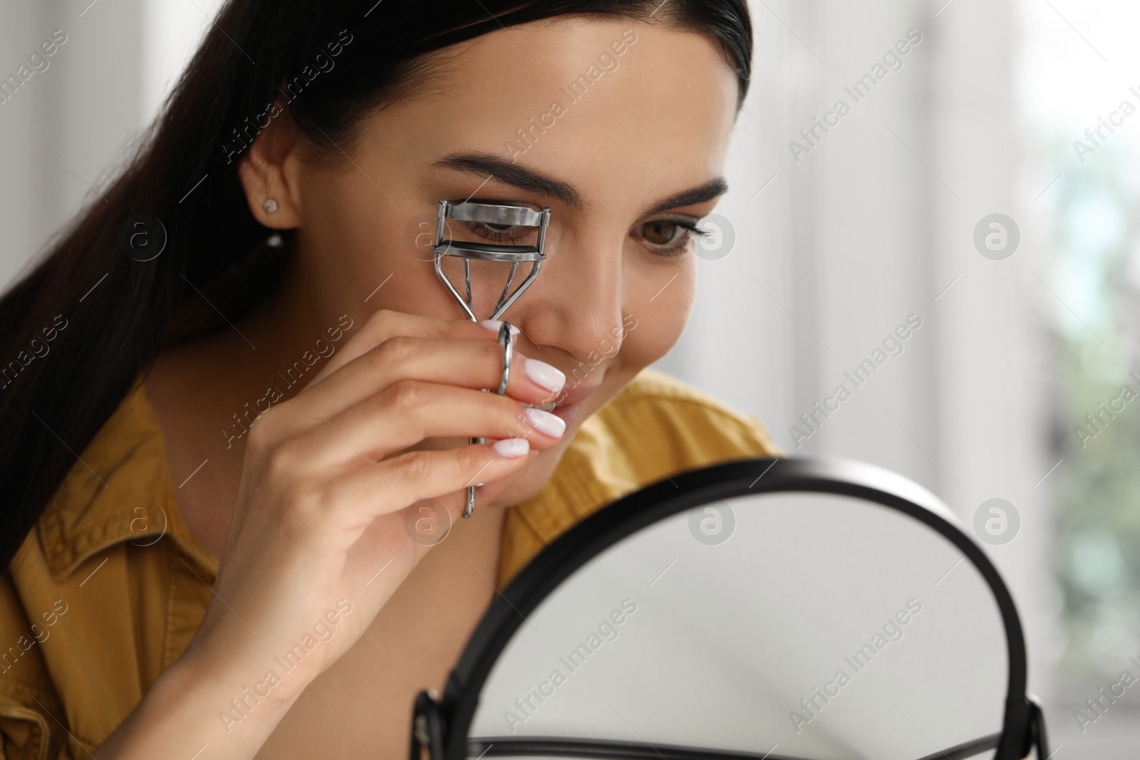 Photo of Beautiful young woman using eyelash curler in front of mirror indoors, closeup