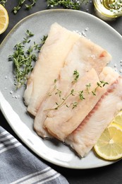 Photo of Plate with raw cod fish, microgreens, lemon and salt on dark table, top view