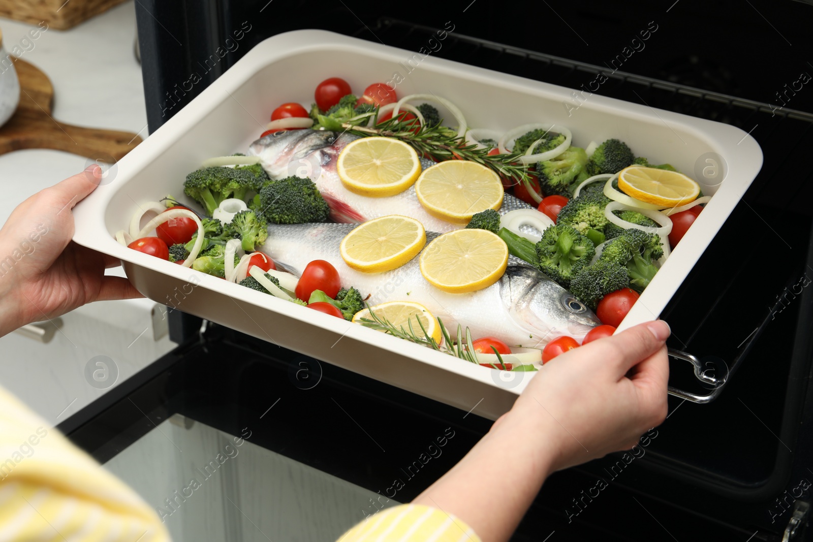 Photo of Woman putting baking dish with raw fish and vegetables into oven in kitchen, closeup