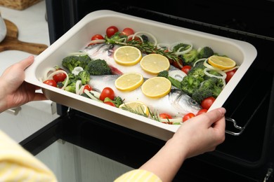 Photo of Woman putting baking dish with raw fish and vegetables into oven in kitchen, closeup