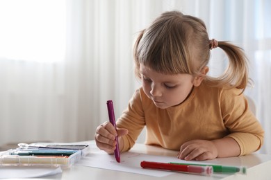 Photo of Cute little girl drawing with marker at white table indoors. Child`s art