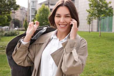 Photo of Woman holding garment cover with clothes while talking on phone outdoors. Dry-cleaning service