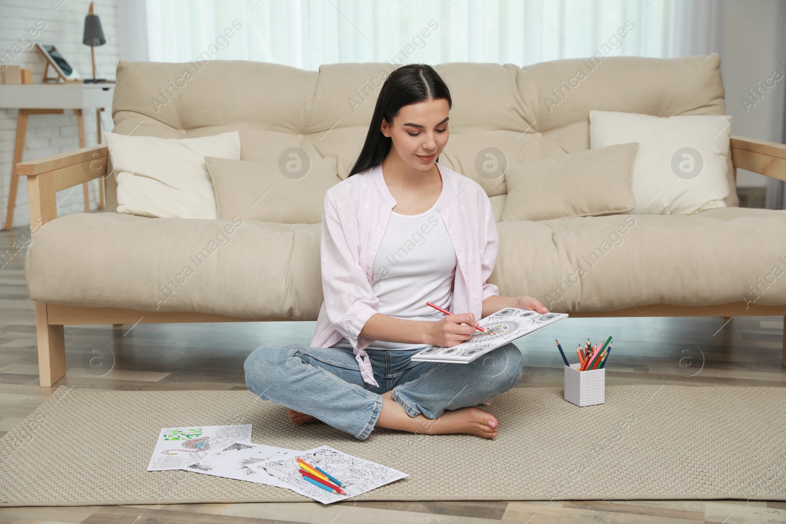 Photo of Young woman coloring antistress page near sofa in living room