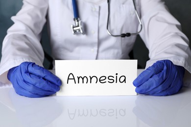 Photo of Doctor holding card with word Amnesia at white table, closeup