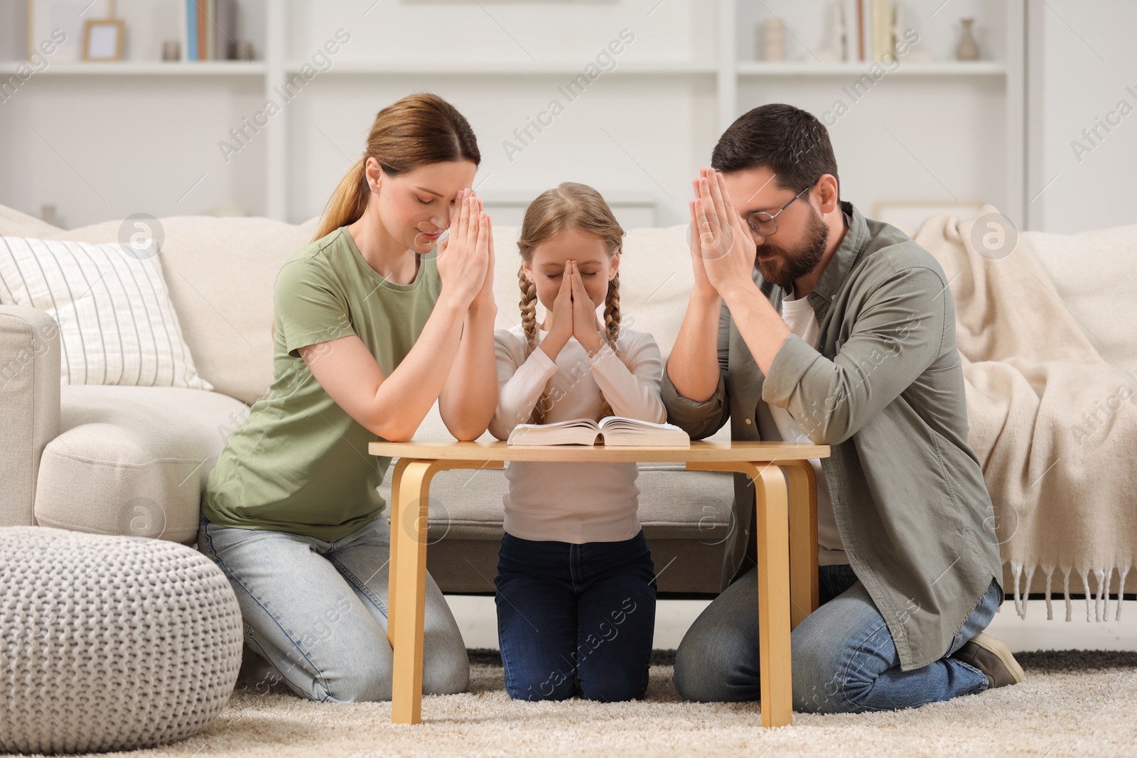 Photo of Girl and her godparents praying over Bible together at table indoors