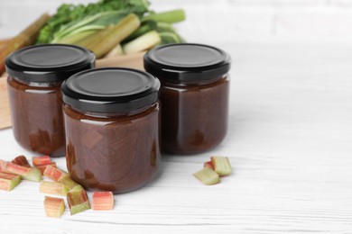Photo of Jars of tasty rhubarb jam and cut stalks on white wooden table, space for text
