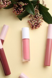 Different lip glosses and flowers on pale yellow background, flat lay