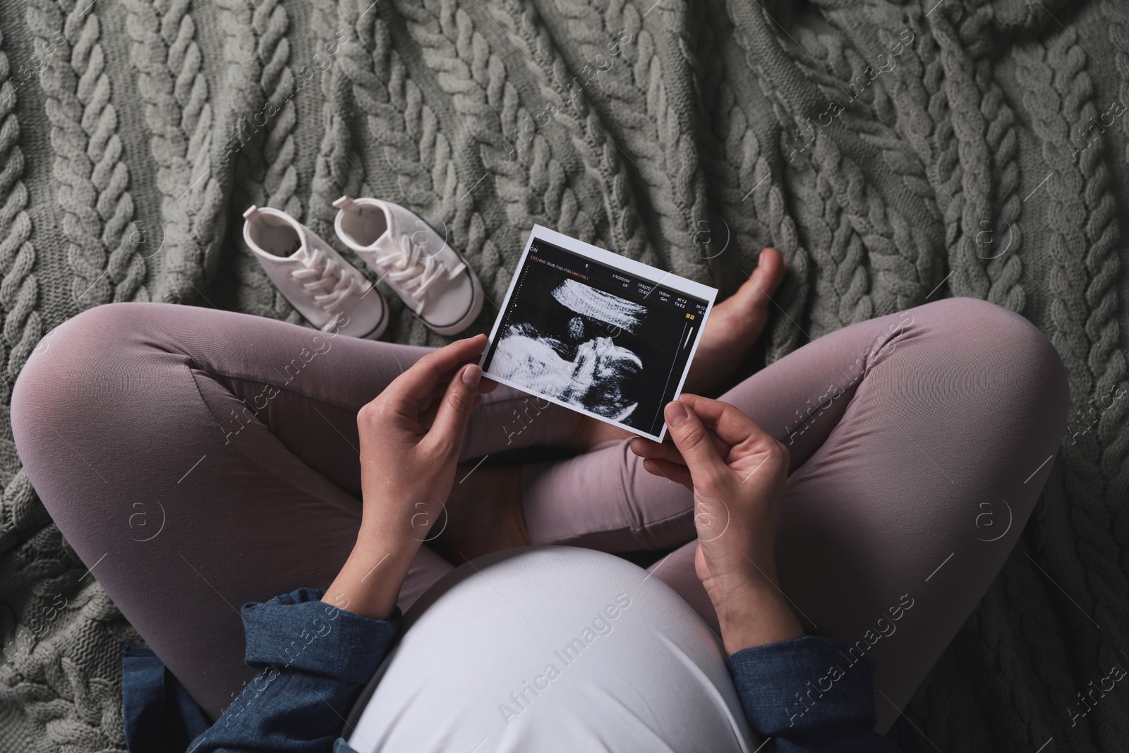 Photo of Young pregnant woman with ultrasound picture of baby and shoes on knitted plaid, top view