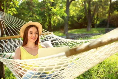 Young woman with laptop resting in comfortable hammock at green garden