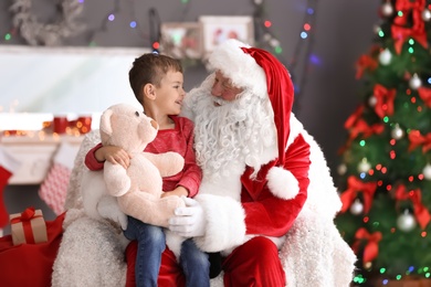 Photo of Little boy with teddy bear sitting on authentic Santa Claus' lap indoors