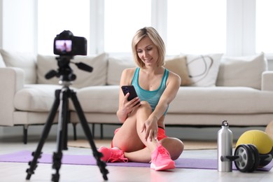 Photo of Smiling sports blogger using smartphone while recording fitness lesson with camera at home