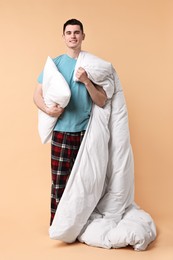 Photo of Happy man in pyjama holding pillow and blanket on beige background