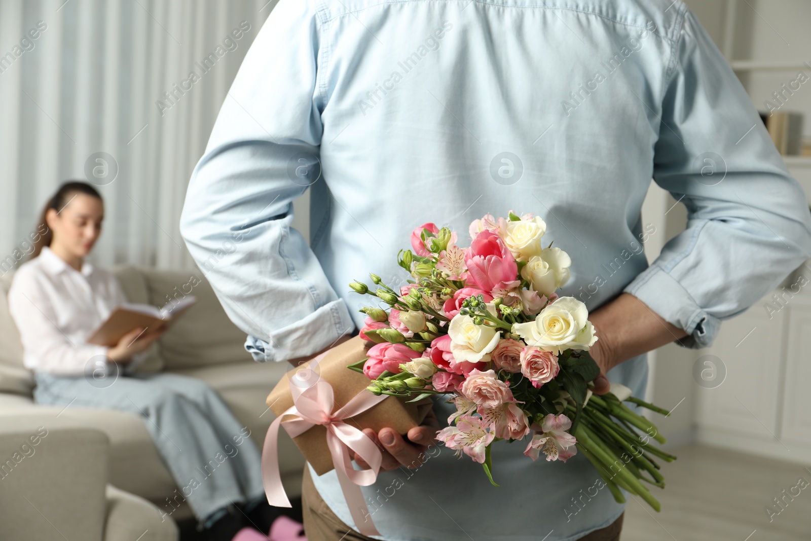 Photo of Man hiding bouquet of flowers and present for his beloved woman indoors, closeup