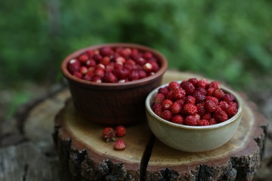 Photo of Bowls of tasty wild strawberries on stump against blurred background, closeup