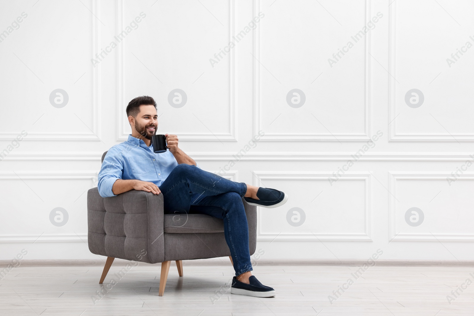 Photo of Handsome man with cup of drink sitting in armchair near white wall indoors, space for text