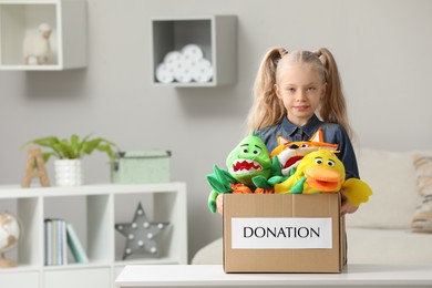 Photo of Cute little girl holding donation box with soft toys at home, space for text