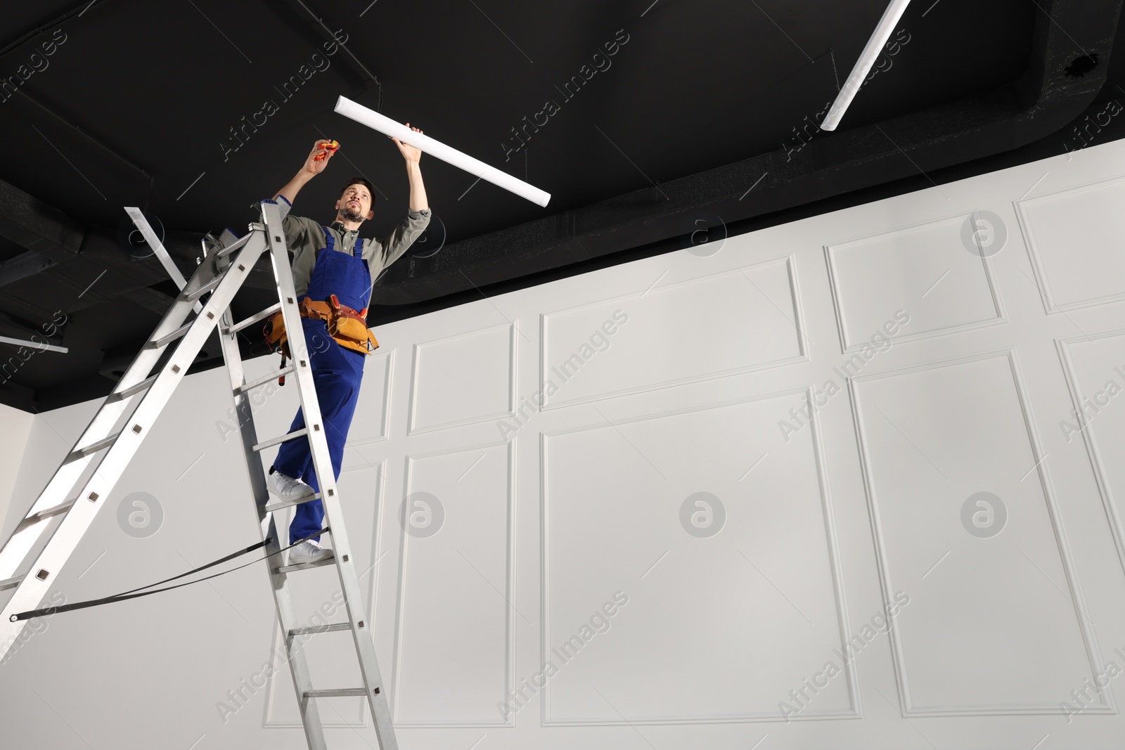 Photo of Electrician in uniform installing ceiling lamp indoors, low angle view