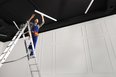 Photo of Electrician in uniform installing ceiling lamp indoors, low angle view