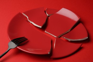 Photo of Pieces of broken ceramic plate and fork on red background, closeup