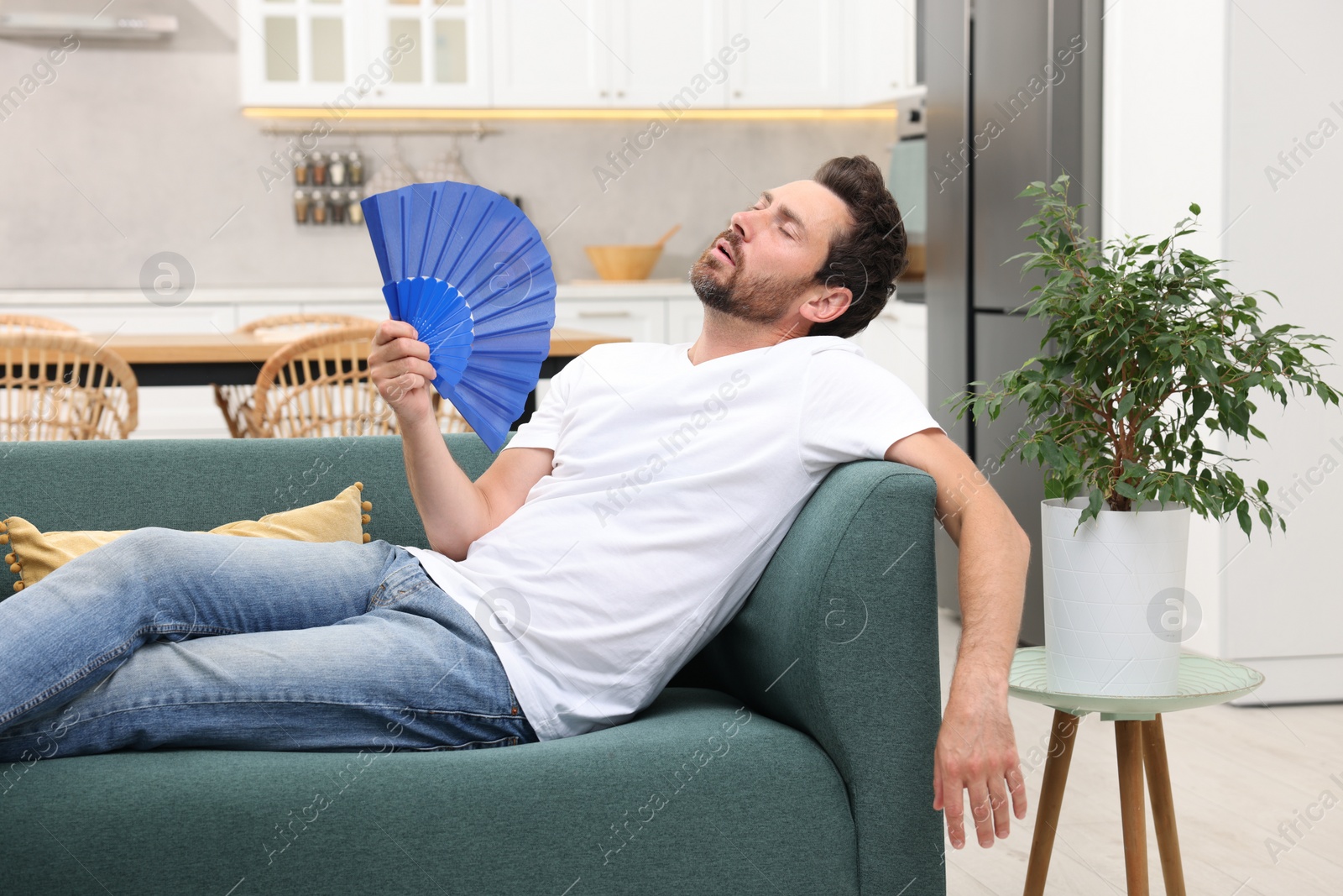 Photo of Bearded man waving blue hand fan to cool himself on sofa at home