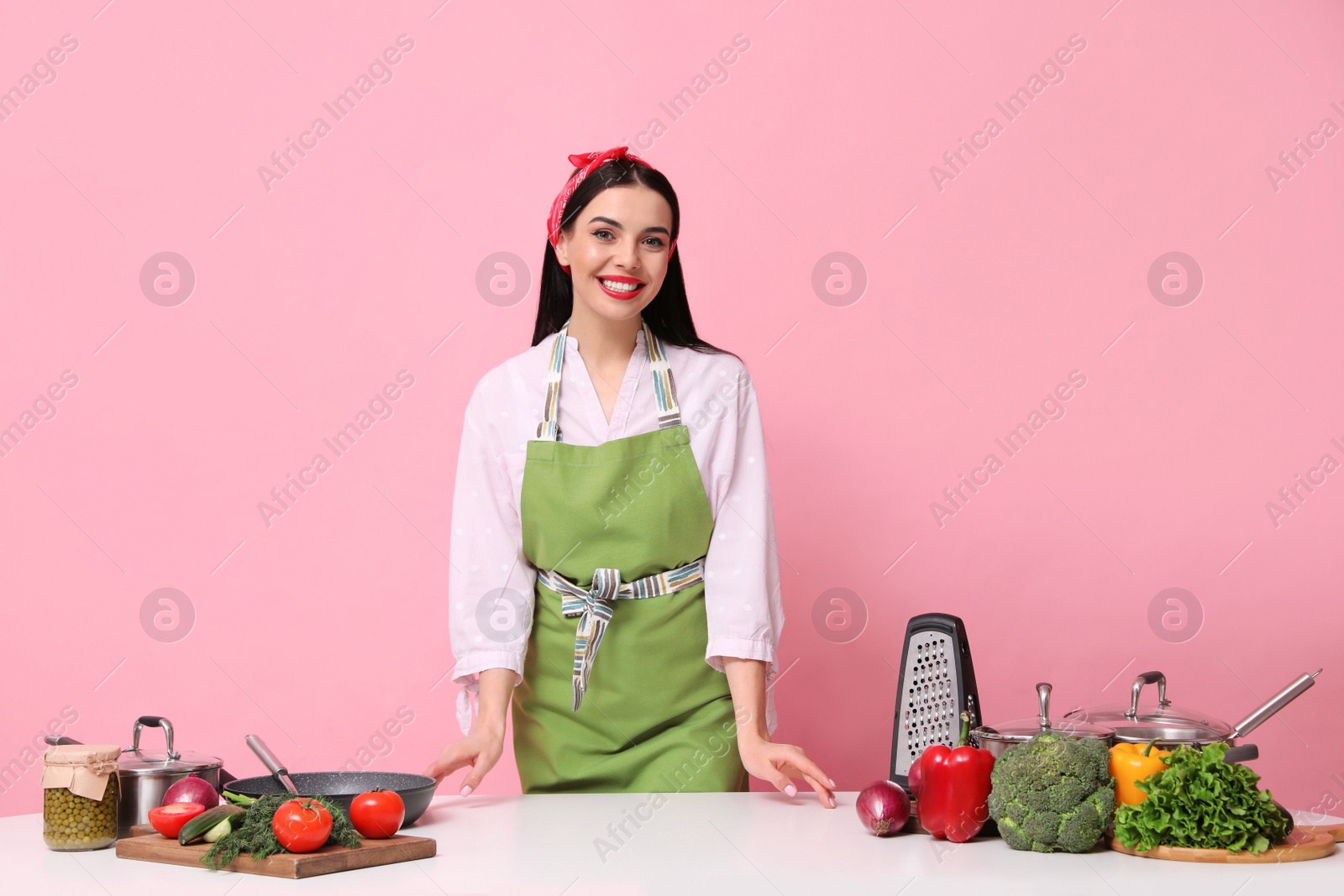 Photo of Young housewife at white table with utensils and products on pink background