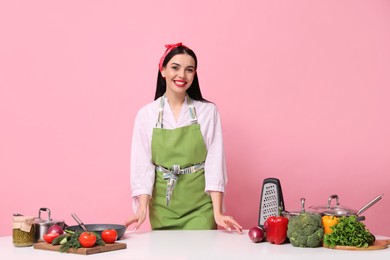Photo of Young housewife at white table with utensils and products on pink background