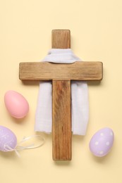 Wooden cross, white cloth and painted Easter eggs on beige background, flat lay