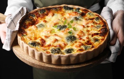 Woman holding wooden board with delicious homemade salmon quiche on black background, closeup