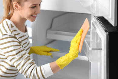 Photo of Woman in rubber gloves cleaning empty refrigerator with rag at home