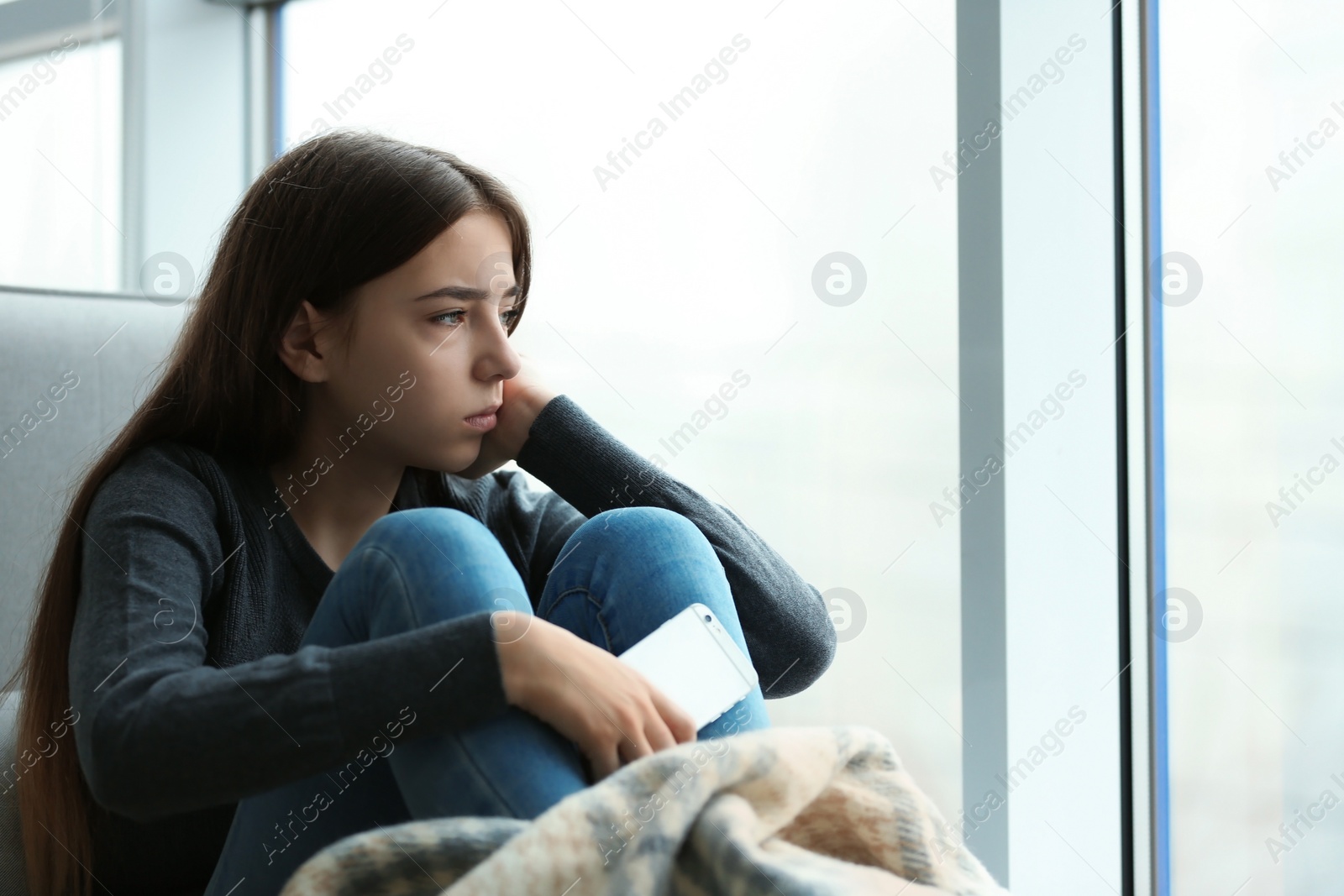 Photo of Upset teenage girl with smartphone sitting at window indoors. Space for text