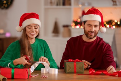Photo of Happy couple in Santa hats decorating Christmas gifts at table in room