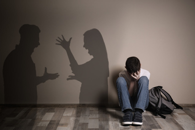 Image of Upset boy with backpack sitting on floor and silhouettes of arguing parents 