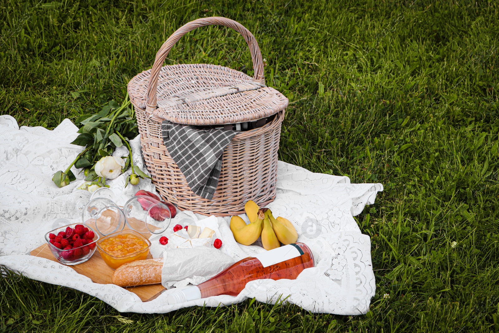 Photo of Picnic blanket with tasty food, flowers, basket and cider on green grass outdoors